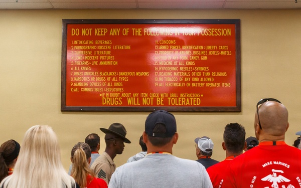 A group of civilians and at least one uniformed military drill instructor stand facing a large red warning sign listing prohibited items at what appears to be a U.S. Marine Corps recruit processing facility. The sign prominently states 'DO NOT KEEP ANY OF THE FOLLOWING IN YOUR POSSESSION' and ends with 'DRUGS WILL NOT BE TOLERATED.' This appears to be a recruit orientation or a civilian visitor tour event at a Marine Corps base.