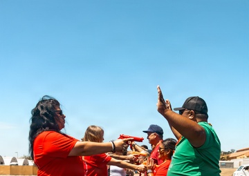 A group of people are engaged in an outdoor team activity or training exercise on a rubber mulch surface under a clear blue sky. A woman in a red shirt is holding a red toy/training gun and pointing it toward another participant in a green shirt who has their hands raised. Several other participants in red shirts are visible in the background, suggesting a coordinated group event.