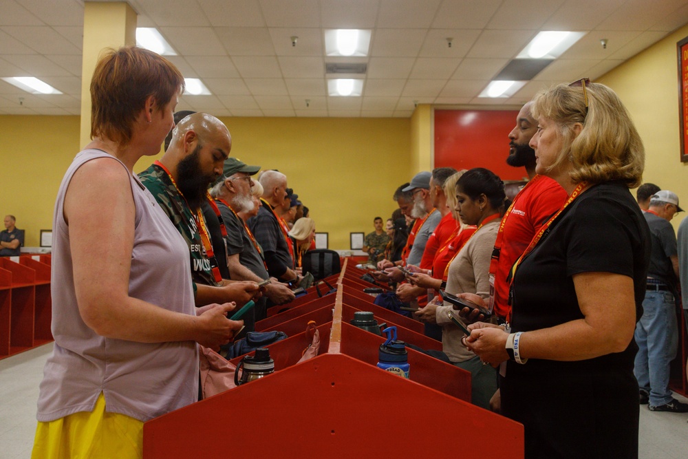 A group of people are gathered in what appears to be an indoor facility with yellow walls, standing on opposite sides of a row of red dividers or partitions. Many participants are wearing orange lanyards and some wear red shirts, suggesting an organized event or program. Participants appear to be looking at their phones or handheld devices.