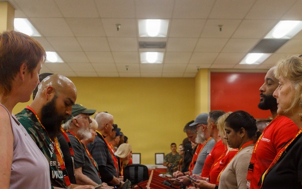 A group of people are gathered in what appears to be an indoor facility with yellow walls, standing on opposite sides of a row of red dividers or partitions. Many participants are wearing orange lanyards and some wear red shirts, suggesting an organized event or program. Participants appear to be looking at their phones or handheld devices.