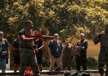 A military demonstration or training exercise is taking place outdoors on a gravel surface, with two Marines in the foreground conducting what appears to be a weapons handling or combat technique demonstration. A group of civilians wearing lanyards and badges observe from the background, suggesting this is a public affairs or community outreach event. Two additional Marines in campaign covers (drill instructor hats) are visible on the right side observing the demonstration.
