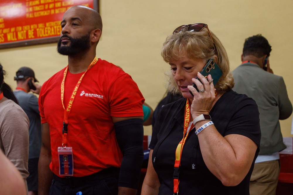 Two individuals wearing Marines-branded lanyards/credentials stand in what appears to be a Marines recruitment or event facility. The man on the left wears a red Marines t-shirt, while the woman on the right is talking on a green smartphone. A yellow wall with a red sign partially visible in the background reads 'WILL NOT BE TOLERATED.'