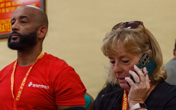 Two individuals wearing Marines-branded lanyards/credentials stand in what appears to be a Marines recruitment or event facility. The man on the left wears a red Marines t-shirt, while the woman on the right is talking on a green smartphone. A yellow wall with a red sign partially visible in the background reads 'WILL NOT BE TOLERATED.'