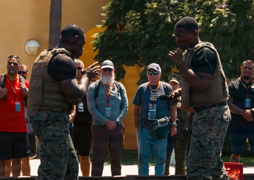 Two U.S. Marines in camouflage uniforms and tactical vests are demonstrating a hand-to-hand combat or martial arts technique on a gravel surface. A group of civilian observers wearing lanyards and badges watch from the perimeter, suggesting this is a demonstration or public affairs event. The setting appears to be an outdoor courtyard of a military installation.