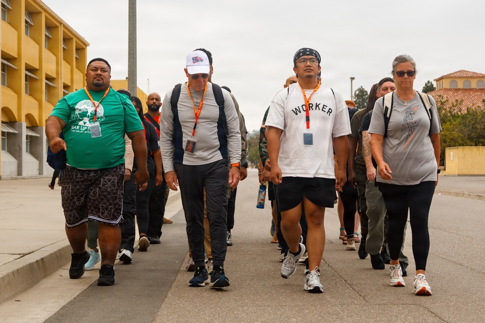 A group of people are walking together along a paved road or pathway, likely participating in an organized walk or event. Several participants are wearing lanyards with badges and carrying backpacks, suggesting a structured group activity. The setting appears to be a institutional or military-style campus based on the surrounding buildings.