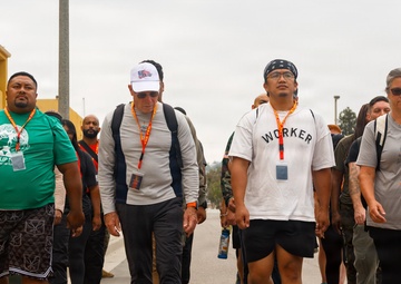 A group of people are walking together along a paved road or pathway, likely participating in an organized walk or event. Several participants are wearing lanyards with badges and carrying backpacks, suggesting a structured group activity. The setting appears to be a institutional or military-style campus based on the surrounding buildings.
