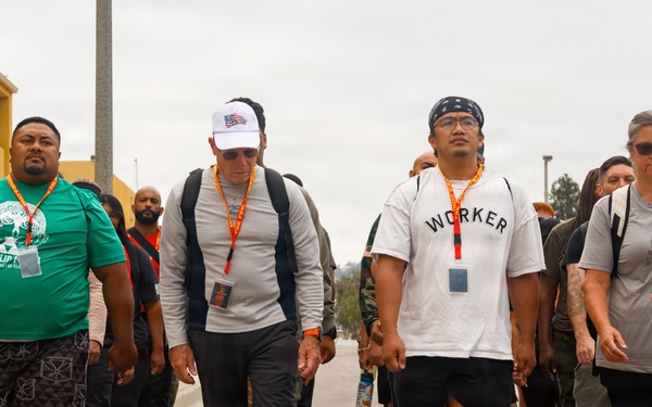 A group of people are walking together along a paved road or pathway, likely participating in an organized walk or event. Several participants are wearing lanyards with badges and carrying backpacks, suggesting a structured group activity. The setting appears to be a institutional or military-style campus based on the surrounding buildings.