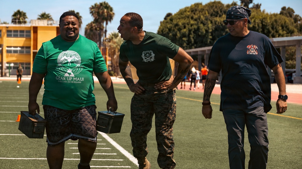 Three men are walking across a football field in what appears to be an outdoor athletic facility. The man on the left is carrying two military-style ammunition cans, wearing a 'Gear Up Maui' green t-shirt, while the center figure is dressed in military camouflage pants and a green uniform shirt. The setting suggests a physical fitness or military training event, with palm trees and school/facility buildings visible in the background.