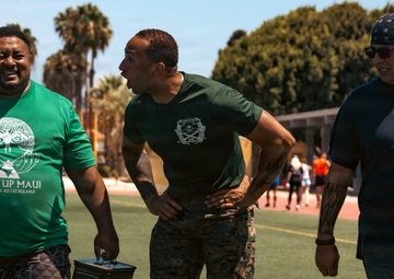 Three men are walking across a football field in what appears to be an outdoor athletic facility. The man on the left is carrying two military-style ammunition cans, wearing a 'Gear Up Maui' green t-shirt, while the center figure is dressed in military camouflage pants and a green uniform shirt. The setting suggests a physical fitness or military training event, with palm trees and school/facility buildings visible in the background.