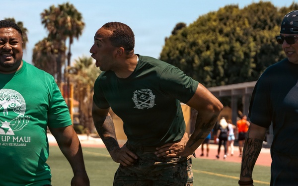Three men are walking across a football field in what appears to be an outdoor athletic facility. The man on the left is carrying two military-style ammunition cans, wearing a 'Gear Up Maui' green t-shirt, while the center figure is dressed in military camouflage pants and a green uniform shirt. The setting suggests a physical fitness or military training event, with palm trees and school/facility buildings visible in the background.