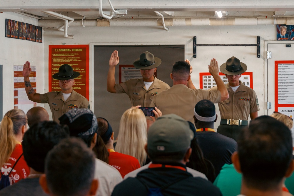 Three United States Marine Corps Drill Instructors in tan service uniforms and campaign covers (drill instructor hats) are raising their right hands, likely reciting an oath or conducting a demonstration for a civilian audience. The scene appears to take place inside a USMC recruiting or training facility, with informational posters visible on the walls. A crowd of civilians, some recording with phones, observes the ceremony from the foreground.