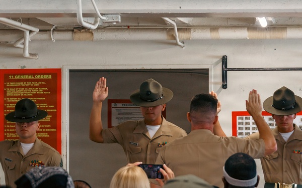 Three United States Marine Corps Drill Instructors in tan service uniforms and campaign covers (drill instructor hats) are raising their right hands, likely reciting an oath or conducting a demonstration for a civilian audience. The scene appears to take place inside a USMC recruiting or training facility, with informational posters visible on the walls. A crowd of civilians, some recording with phones, observes the ceremony from the foreground.