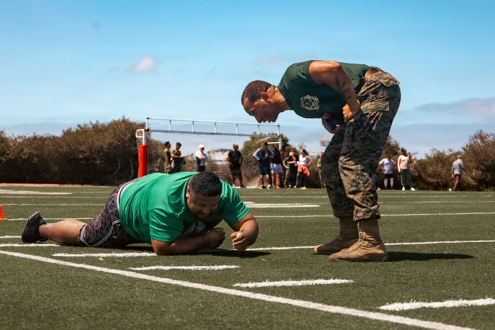 A military instructor in Marine Corps camouflage utilities and a green t-shirt leans over and appears to be motivating or instructing a civilian in a green shirt who is lying prone on a turf athletic field. The scene appears to be a fitness assessment or physical training event, likely a recruiting or community outreach activity. A group of spectators is visible in the background near a soccer goal.