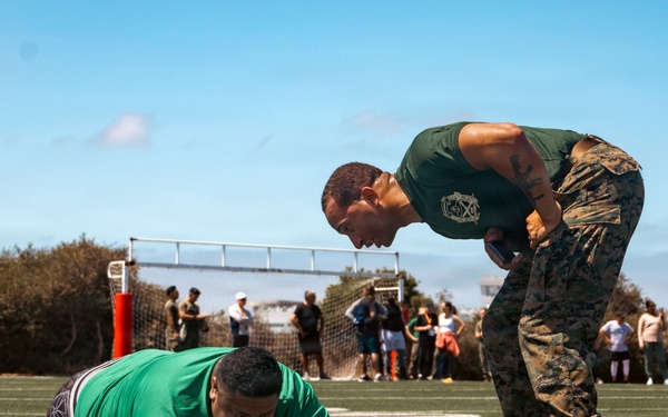 A military instructor in Marine Corps camouflage utilities and a green t-shirt leans over and appears to be motivating or instructing a civilian in a green shirt who is lying prone on a turf athletic field. The scene appears to be a fitness assessment or physical training event, likely a recruiting or community outreach activity. A group of spectators is visible in the background near a soccer goal.