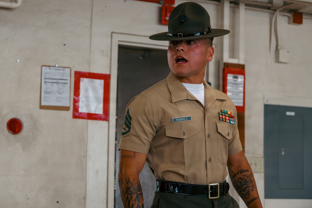 A United States Marine Corps Drill Instructor named Ramirez is photographed in what appears to be a barracks or training facility. He is wearing the distinctive campaign cover (drill instructor hat), khaki service uniform with rank insignia and service ribbons, and has visible tattoos on both forearms. His mouth is open, suggesting he is issuing commands or instructions.