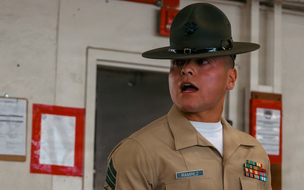 A United States Marine Corps Drill Instructor named Ramirez is photographed in what appears to be a barracks or training facility. He is wearing the distinctive campaign cover (drill instructor hat), khaki service uniform with rank insignia and service ribbons, and has visible tattoos on both forearms. His mouth is open, suggesting he is issuing commands or instructions.