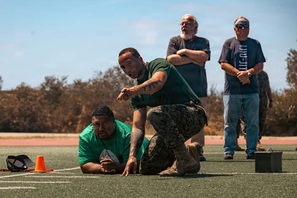 A military or law enforcement training exercise is taking place on an outdoor athletic field. One instructor in camouflage pants and a green shirt is kneeling over a participant lying prone on the ground, pointing and giving directions. Two older civilian observers stand in the background watching the demonstration.