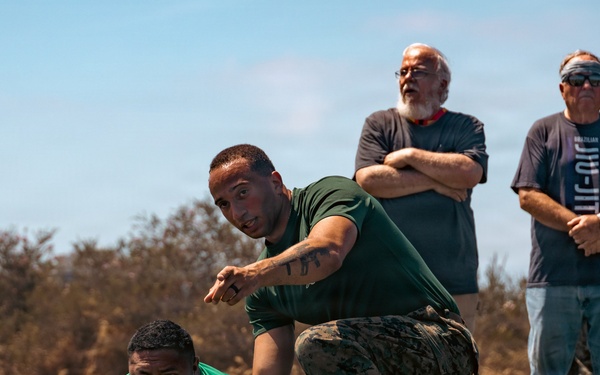 A military or law enforcement training exercise is taking place on an outdoor athletic field. One instructor in camouflage pants and a green shirt is kneeling over a participant lying prone on the ground, pointing and giving directions. Two older civilian observers stand in the background watching the demonstration.