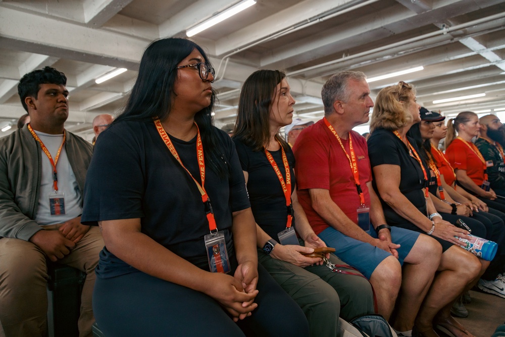 A group of people are seated in rows, attentively watching or listening to something off-camera in what appears to be an indoor venue with industrial-style fluorescent lighting. Most attendees are wearing orange lanyards with visible text reading 'THE FEW' and 'MARINES,' suggesting this is an organized military or recruitment-related event. The audience appears engaged and focused, dressed casually in dark shirts and civilian clothing.