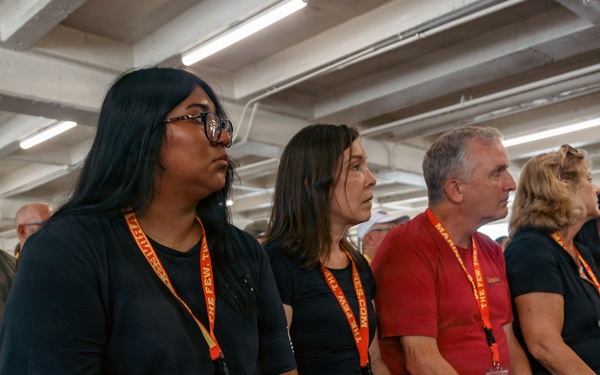 A group of people are seated in rows, attentively watching or listening to something off-camera in what appears to be an indoor venue with industrial-style fluorescent lighting. Most attendees are wearing orange lanyards with visible text reading 'THE FEW' and 'MARINES,' suggesting this is an organized military or recruitment-related event. The audience appears engaged and focused, dressed casually in dark shirts and civilian clothing.