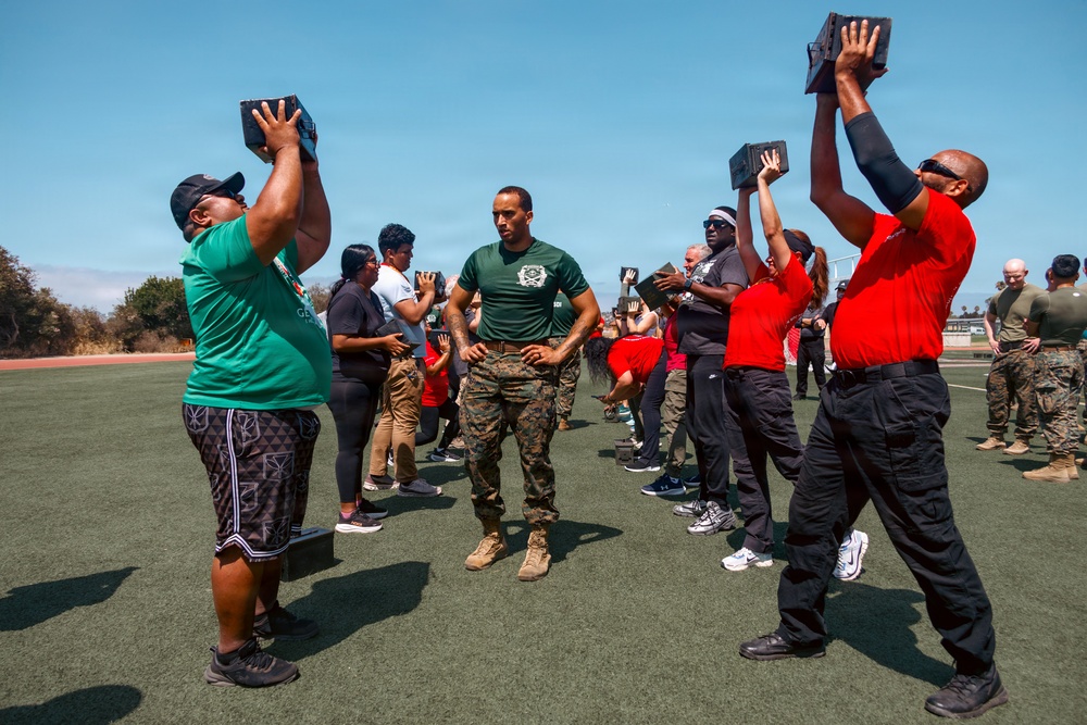 A military instructor in camouflage uniform and green t-shirt oversees a group of civilians performing an overhead exercise with heavy black blocks on an outdoor athletic field. The participants are wearing casual clothing in red and green, suggesting this may be a community outreach or civilian-military fitness event. Marines in uniform can be seen observing in the background.