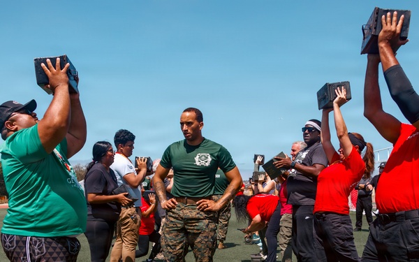 A military instructor in camouflage uniform and green t-shirt oversees a group of civilians performing an overhead exercise with heavy black blocks on an outdoor athletic field. The participants are wearing casual clothing in red and green, suggesting this may be a community outreach or civilian-military fitness event. Marines in uniform can be seen observing in the background.