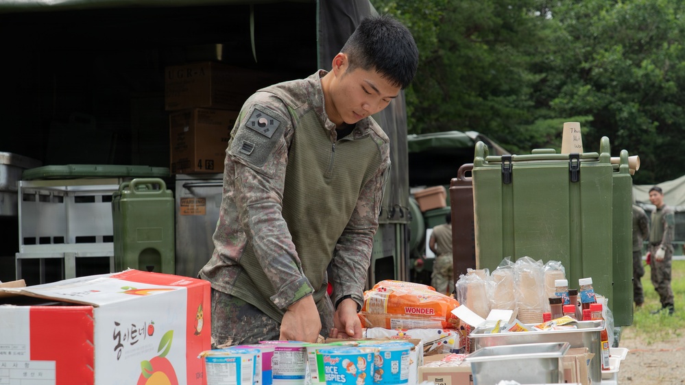 A soldier in camouflage uniform, bearing a South Korean flag patch on the sleeve, is organizing food supplies on a table at what appears to be a military field camp. The table contains a variety of food items including American and Korean branded products. The setting is an outdoor military logistics or field kitchen area with military vehicles and equipment visible in the background.