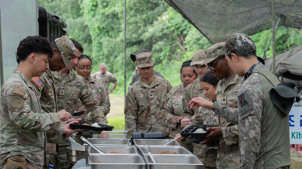 A group of U.S. Army soldiers in MultiCam/OCP camouflage uniforms are going through a field chow line at an outdoor military field kitchen. They are serving themselves food from stainless steel serving trays onto black meal trays. The setting appears to be a forward operating or training area with green vegetation and military tentage visible in the background.