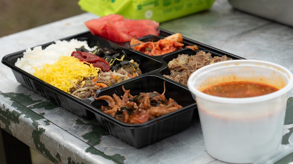A Korean-style bento box (dosirak) is displayed on a weathered outdoor table, containing multiple compartments filled with various side dishes (banchan), rice, and a main protein. Alongside the tray sits a white styrofoam cup filled with a red, spicy-looking soup or broth. The meal appears to be a takeout or outdoor dining setup, typical of Korean casual food service.