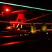 A nighttime scene on an aircraft carrier flight deck showing deck crew personnel and at least one fixed-wing aircraft, illuminated by red and green light streaks consistent with a long-exposure photograph. The image captures the dynamic, high-intensity environment of carrier flight operations after dark. Light trails suggest movement of aircraft or deck equipment during the exposure.