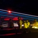 A long-exposure night photograph taken on what appears to be an aircraft carrier flight deck, capturing light trails from aircraft navigation lights and deck equipment. Several flight deck crew members are visible in silhouette, wearing helmets and flight deck gear. The image combines motion blur of moving lights with static elements to create a dramatic visual effect.