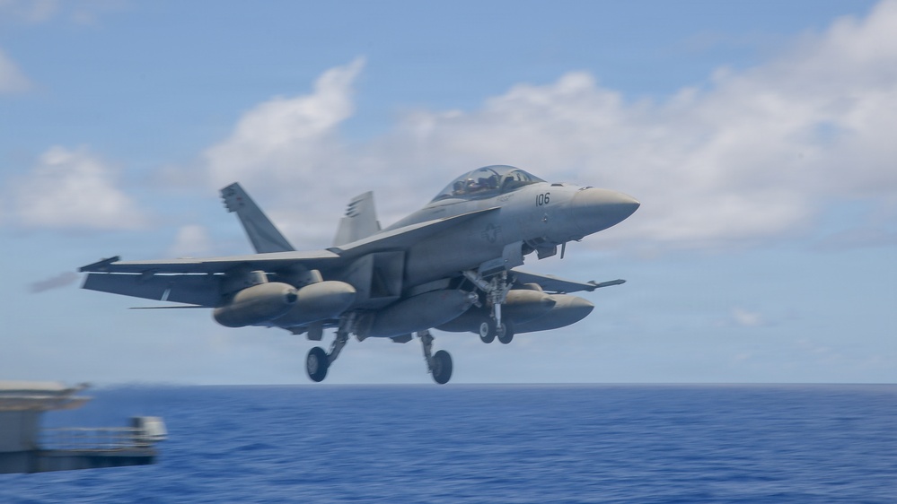 A U.S. Navy F/A-18 Super Hornet (bureau number 106) is captured mid-launch or low-altitude flight just off the deck of an aircraft carrier over open ocean. The aircraft has its landing gear fully extended and is carrying external fuel tanks and ordnance on its hardpoints. The image was taken from the carrier deck level, providing a close perspective of the jet against a partly cloudy sky.