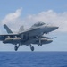 A U.S. Navy F/A-18 Super Hornet (bureau number 106) is captured mid-launch or low-altitude flight just off the deck of an aircraft carrier over open ocean. The aircraft has its landing gear fully extended and is carrying external fuel tanks and ordnance on its hardpoints. The image was taken from the carrier deck level, providing a close perspective of the jet against a partly cloudy sky.