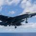 A U.S. Navy carrier-based jet fighter, marked with the number '410', is photographed in flight with landing gear deployed, suggesting an approach or recent launch over open ocean. The aircraft appears to be an F/A-18E/F Super Hornet in a dark tactical paint scheme. The background shows a partly cloudy sky over a deep blue ocean horizon.