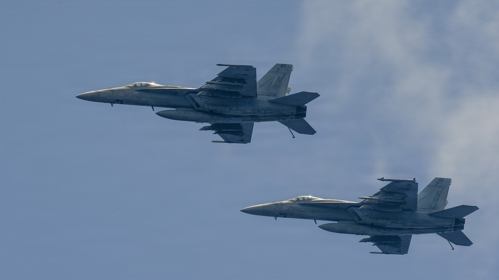 Two military jet fighters are flying in formation against a partly cloudy blue sky. Both aircraft appear to be F/A-18 Super Hornets, identifiable by their twin tails, twin engines, and distinctive airframe shape. The aircraft are flying in a loose trail formation, with one aircraft positioned slightly ahead and above the other.