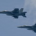 Two military jet fighters are flying in formation against a partly cloudy blue sky. Both aircraft appear to be F/A-18 Super Hornets, identifiable by their twin tails, twin engines, and distinctive airframe shape. The aircraft are flying in a loose trail formation, with one aircraft positioned slightly ahead and above the other.