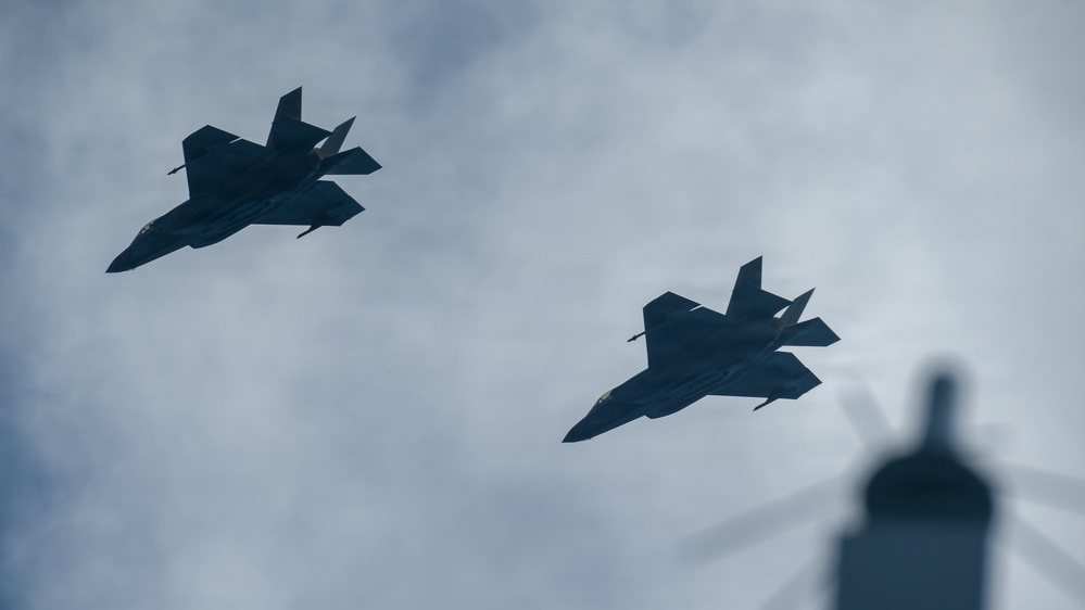 Two military stealth fighter jets are captured in silhouette flying in formation against an overcast sky. The aircraft display angular, low-observable design characteristics consistent with fifth-generation stealth fighters. A blurred structure, possibly a tower or antenna, is partially visible in the lower right corner.