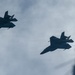 Two military stealth fighter jets are captured in silhouette flying in formation against an overcast sky. The aircraft display angular, low-observable design characteristics consistent with fifth-generation stealth fighters. A blurred structure, possibly a tower or antenna, is partially visible in the lower right corner.