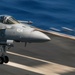 A U.S. Navy F/A-18 Super Hornet marked '404' is captured during a carrier launch or landing on an aircraft carrier flight deck. The aircraft is at low altitude above the deck with landing gear deployed, set against a deep blue ocean background. Motion blur on the deck surface indicates high speed and dynamic action.