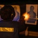 A person is firing a handgun at a shooting range during nighttime or low-light conditions. The shooter is facing away from the camera, aiming at standard silhouette paper targets. A muzzle flash is clearly visible, illuminating the immediate area around the firearm.