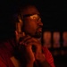 A man is photographed in a very dark environment, illuminated primarily by warm orange/red light from the left side. He is wearing protective eyewear and over-ear hearing protection, holding what appears to be a firearm raised near his face in a shooting stance. The background shows blurred rectangular light panels, suggesting an indoor shooting range environment.