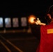 A person wearing a red jacket and ear protection is firing a handgun at a shooting range during nighttime or low-light conditions. A visible muzzle flash is captured at the moment of discharge. Multiple illuminated paper targets are visible downrange in the background.