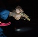 A person is handling a tan/flat dark earth colored semi-automatic pistol in a dark environment. The individual is wearing a blue shirt and red garment, with a gold chain bracelet visible on their wrist. The image appears to be taken at a shooting range or similar low-light setting.