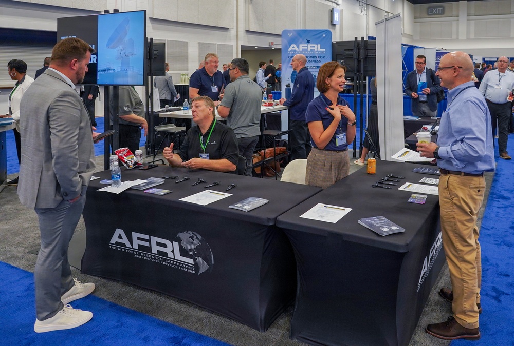 This image shows an AFRL (Air Force Research Laboratory) booth at what appears to be a business or defense industry trade show or conference. Representatives are engaged in conversations with attendees at a black-draped table displaying brochures, pens, and informational materials. A banner in the background identifies the booth as the AFRL Small Business Office with the tagline 'Open Doors for Business.'
