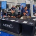 This image shows an AFRL (Air Force Research Laboratory) booth at what appears to be a business or defense industry trade show or conference. Representatives are engaged in conversations with attendees at a black-draped table displaying brochures, pens, and informational materials. A banner in the background identifies the booth as the AFRL Small Business Office with the tagline 'Open Doors for Business.'