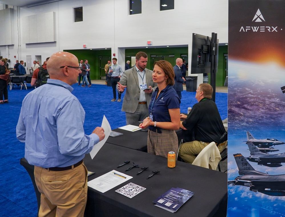 An AFWERX booth at what appears to be a defense or aerospace industry conference or trade show. A woman in a navy AFWERX polo shirt is engaging with attendees at a black-draped table, while a seated colleague and a standing man in a suit also participate. The booth features AFWERX branding, a banner displaying F-16 fighter jets, a QR code, printed materials, and a monitor.