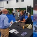 An AFWERX booth at what appears to be a defense or aerospace industry conference or trade show. A woman in a navy AFWERX polo shirt is engaging with attendees at a black-draped table, while a seated colleague and a standing man in a suit also participate. The booth features AFWERX branding, a banner displaying F-16 fighter jets, a QR code, printed materials, and a monitor.