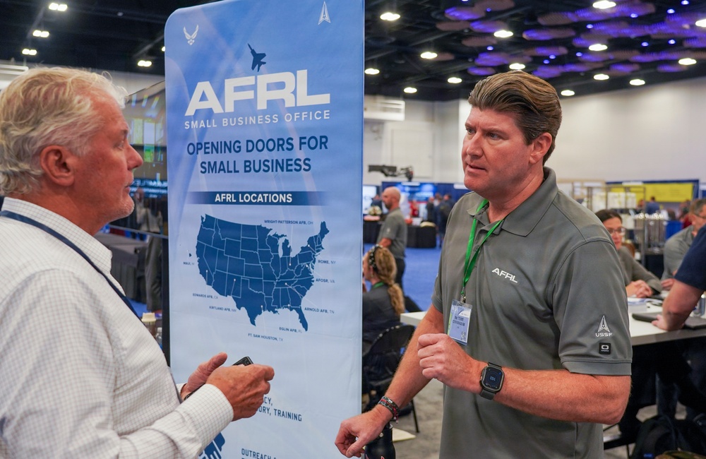 Two men are engaged in conversation at a conference or trade show booth for the AFRL (Air Force Research Laboratory) Small Business Office. The booth displays a banner reading 'Opening Doors for Small Business' with a map showing AFRL locations across the United States. The man on the right is wearing an AFRL-branded polo shirt with a USSF (United States Space Force) logo, suggesting he is an AFRL representative.
