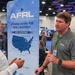 Two men are engaged in conversation at a conference or trade show booth for the AFRL (Air Force Research Laboratory) Small Business Office. The booth displays a banner reading 'Opening Doors for Small Business' with a map showing AFRL locations across the United States. The man on the right is wearing an AFRL-branded polo shirt with a USSF (United States Space Force) logo, suggesting he is an AFRL representative.