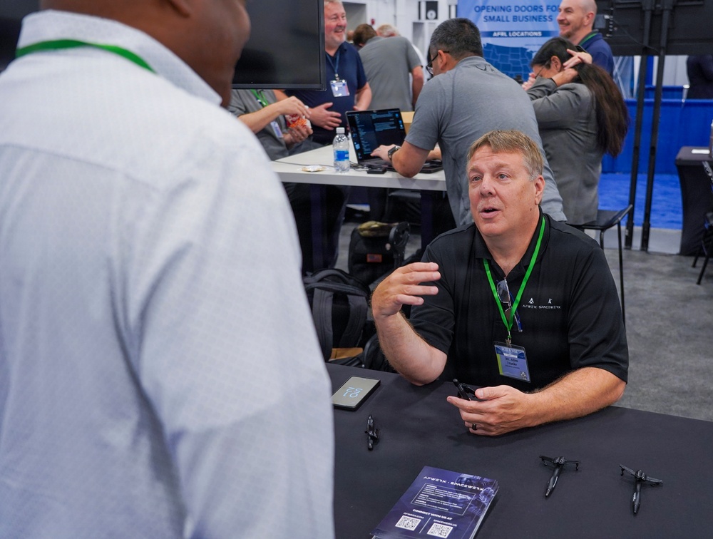 A professional trade show or conference event where attendees are networking and engaging in business discussions. A man in a black shirt with an 'Apex Spaceworks' logo and green lanyard is seated at a booth table, actively conversing with another attendee. The background shows additional booth activity with a sign reading 'Opening Doors for Small Business' and multiple people interacting.